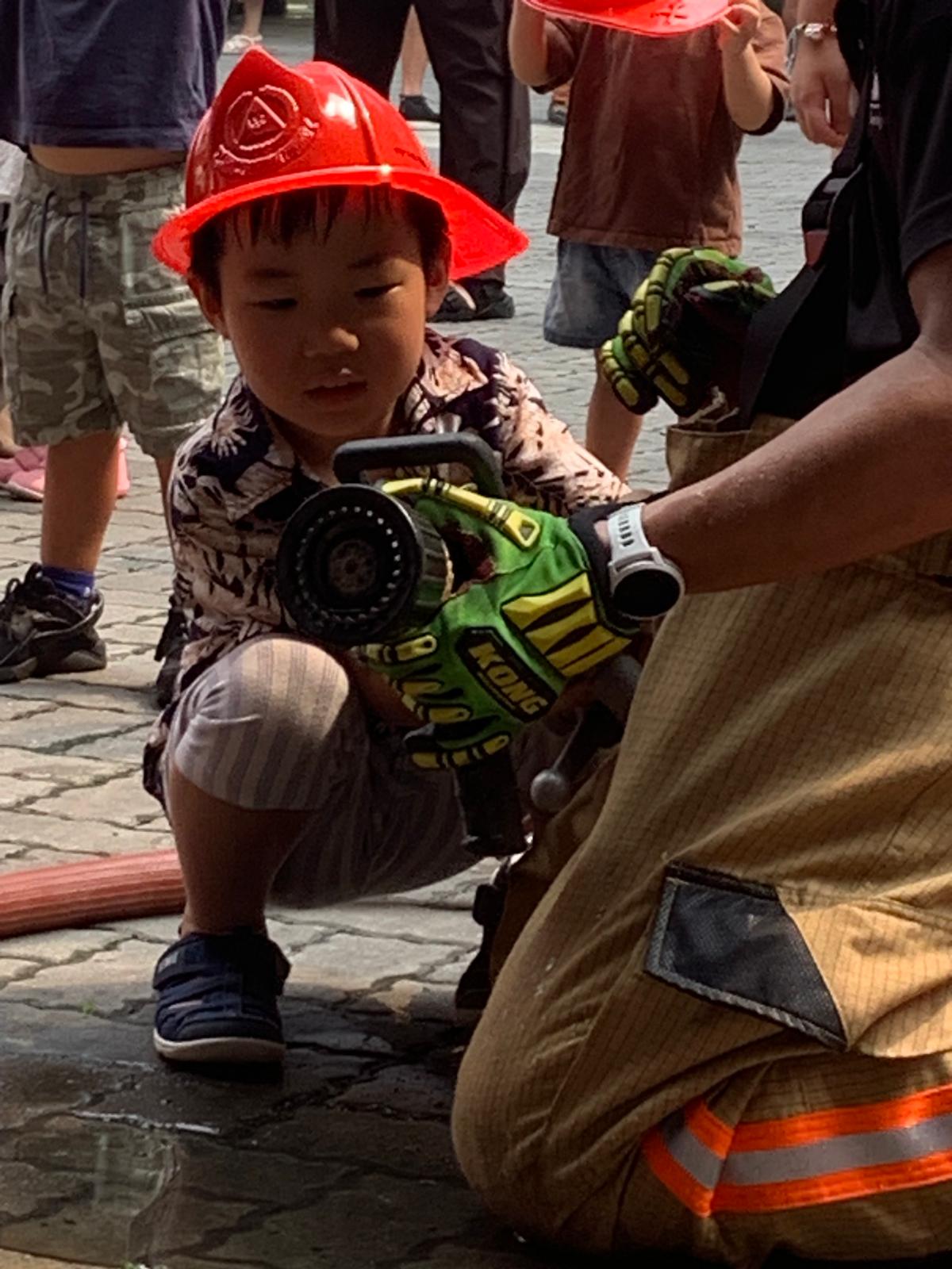 Child in a red helmet exploring a fire hose with a firefighter